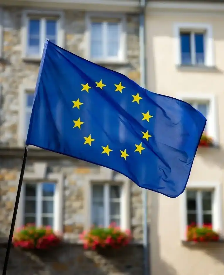 Drapeau de l’Union européenne bleu avec douze étoiles dorées flottant devant une façade en pierre et fenêtres fleuries, symbole d’unité et de paix en Europe.
